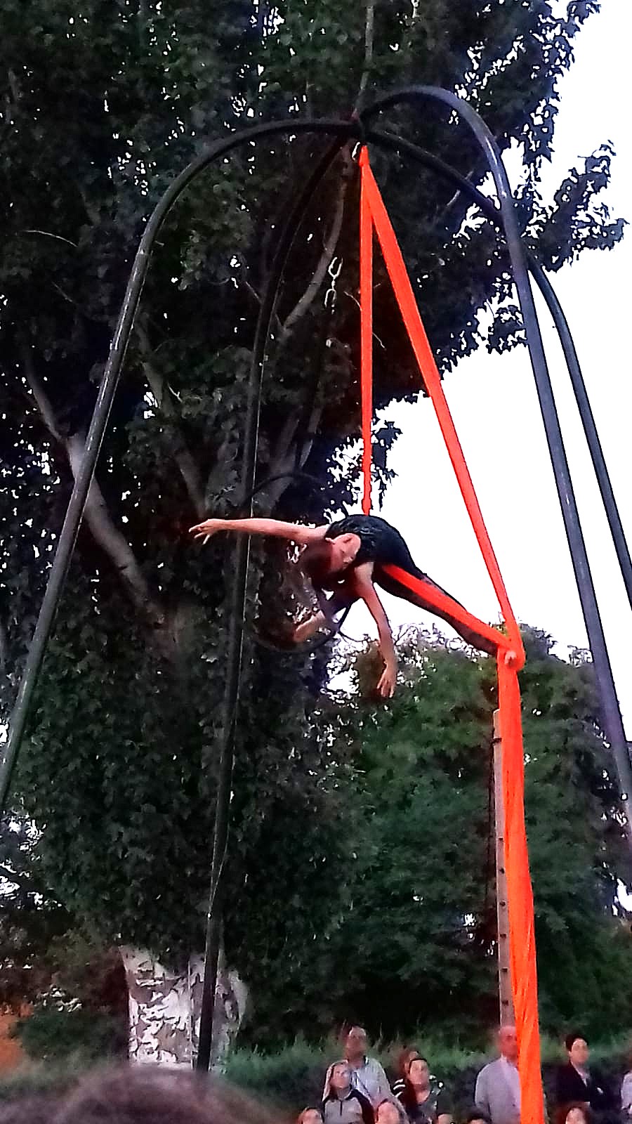 Acróbata aérea suspendida en telas naranjas desde un pórtico metálico.