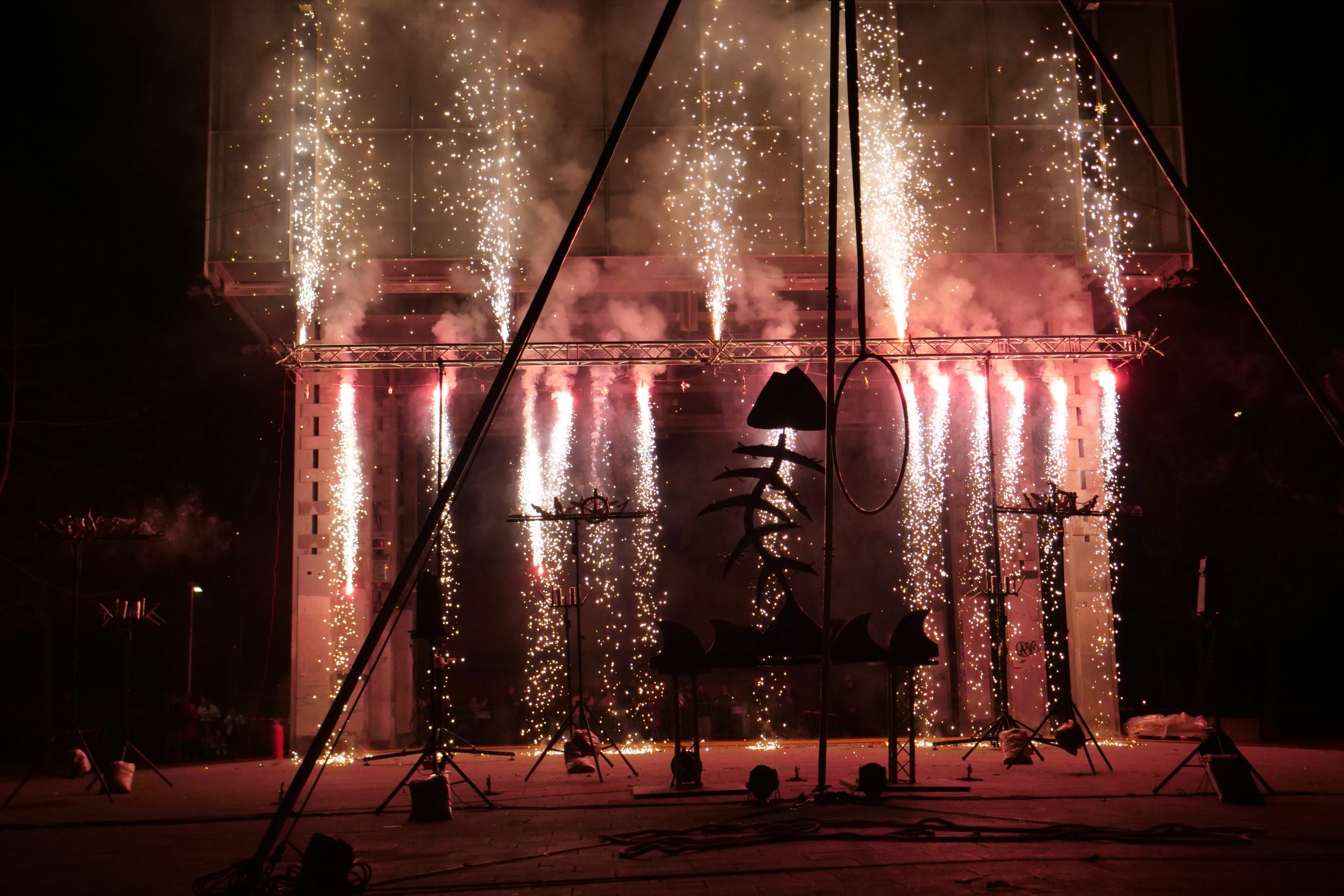Columnas de chispas blancas y rosadas caen desde una estructura metálica, creando una cortina de fuego y luz.
