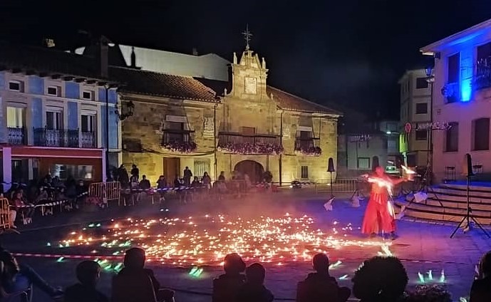 Un artista de rojo danza en un escenario circular de fuego frente a edificios antiguos.