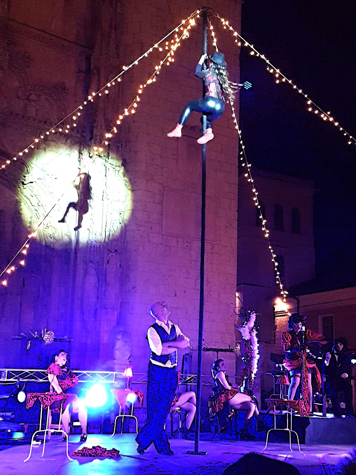 Una acróbata con mallas brillantes trepa por un mástil vertical rodeado de guirnaldas de luces.