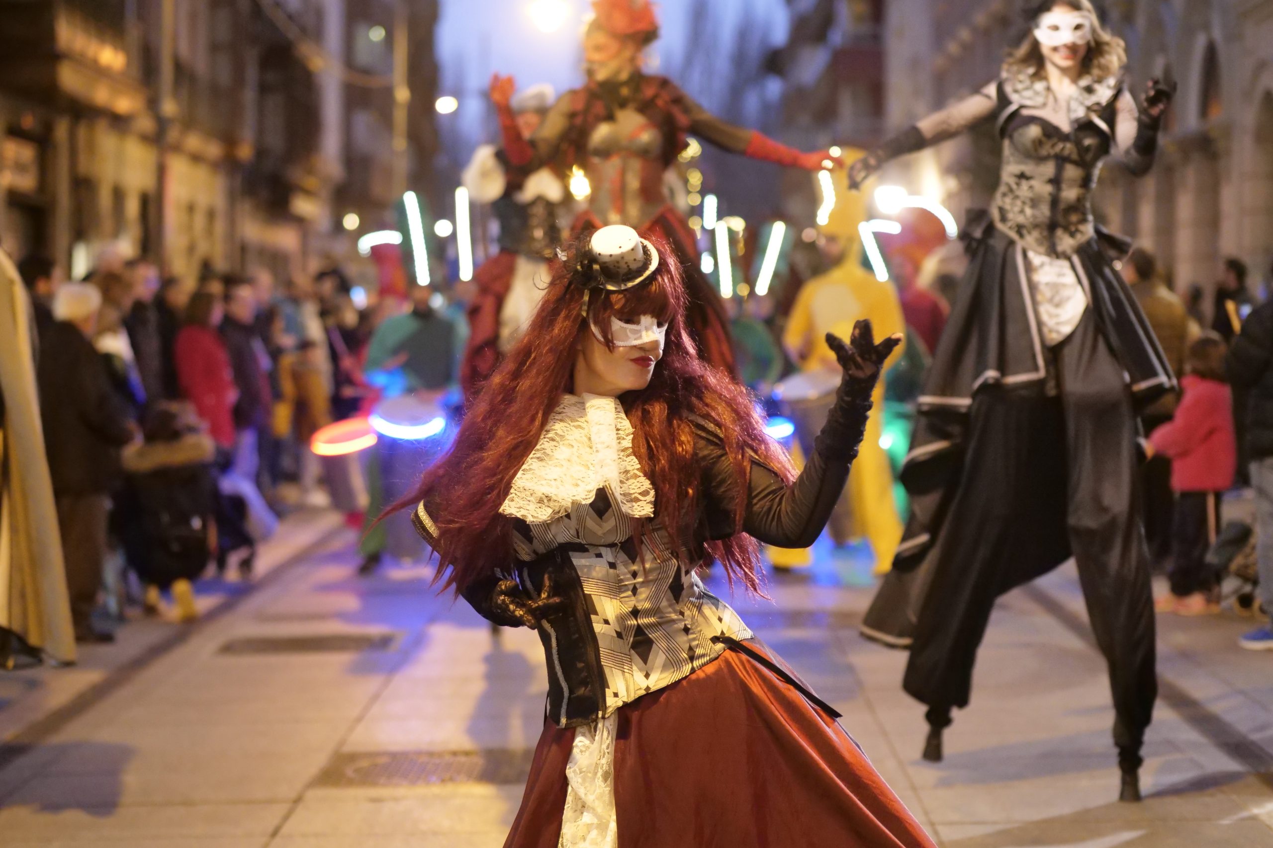 Desfile nocturno con artistas de estética carnavalesca. una bailarina con antifaz blanco y melena cobriza danza en primer plano.