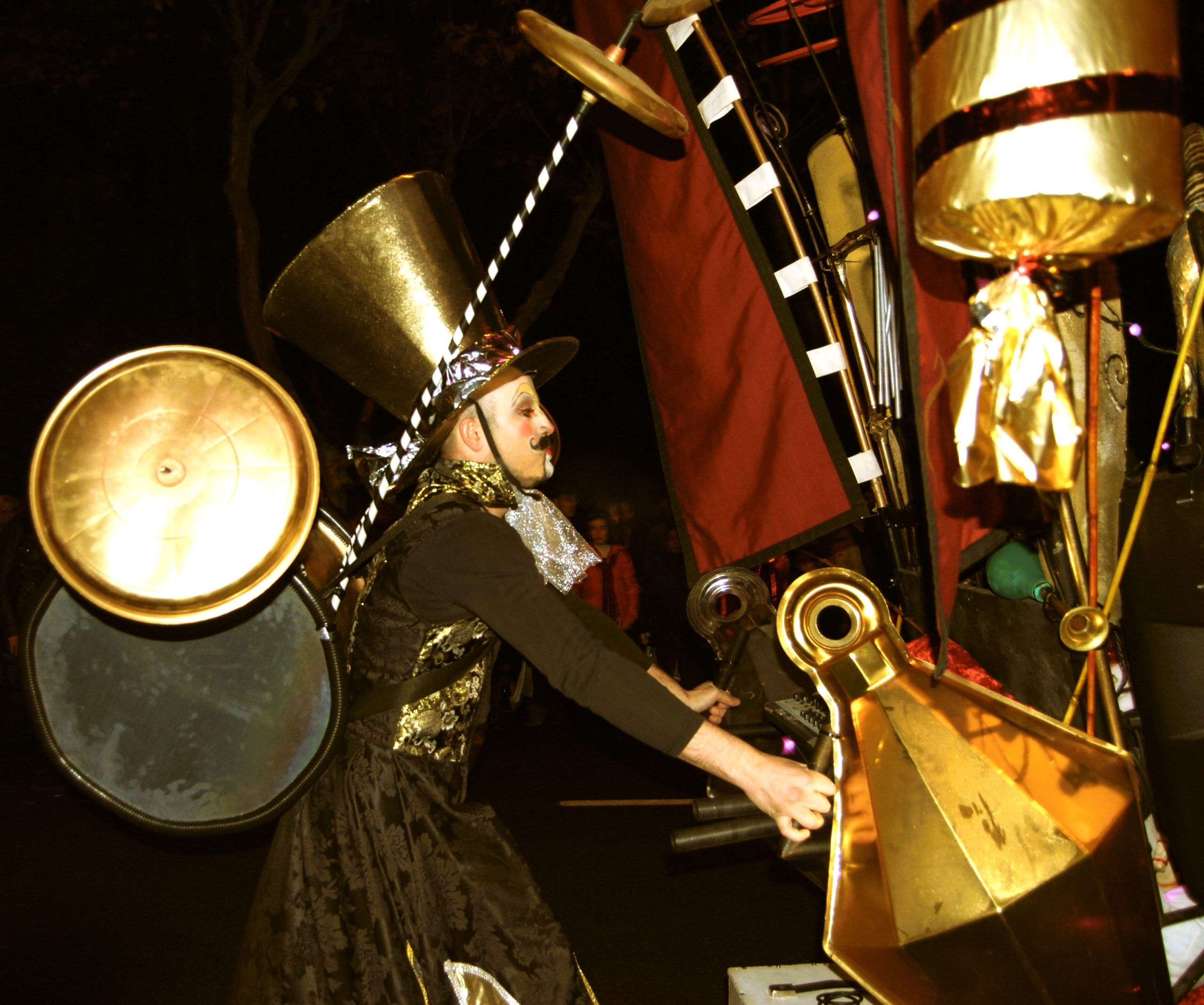 Artista con un traje dorado y negro y un sombrero de copa gigante maneja una estructura de percusión móvil.
