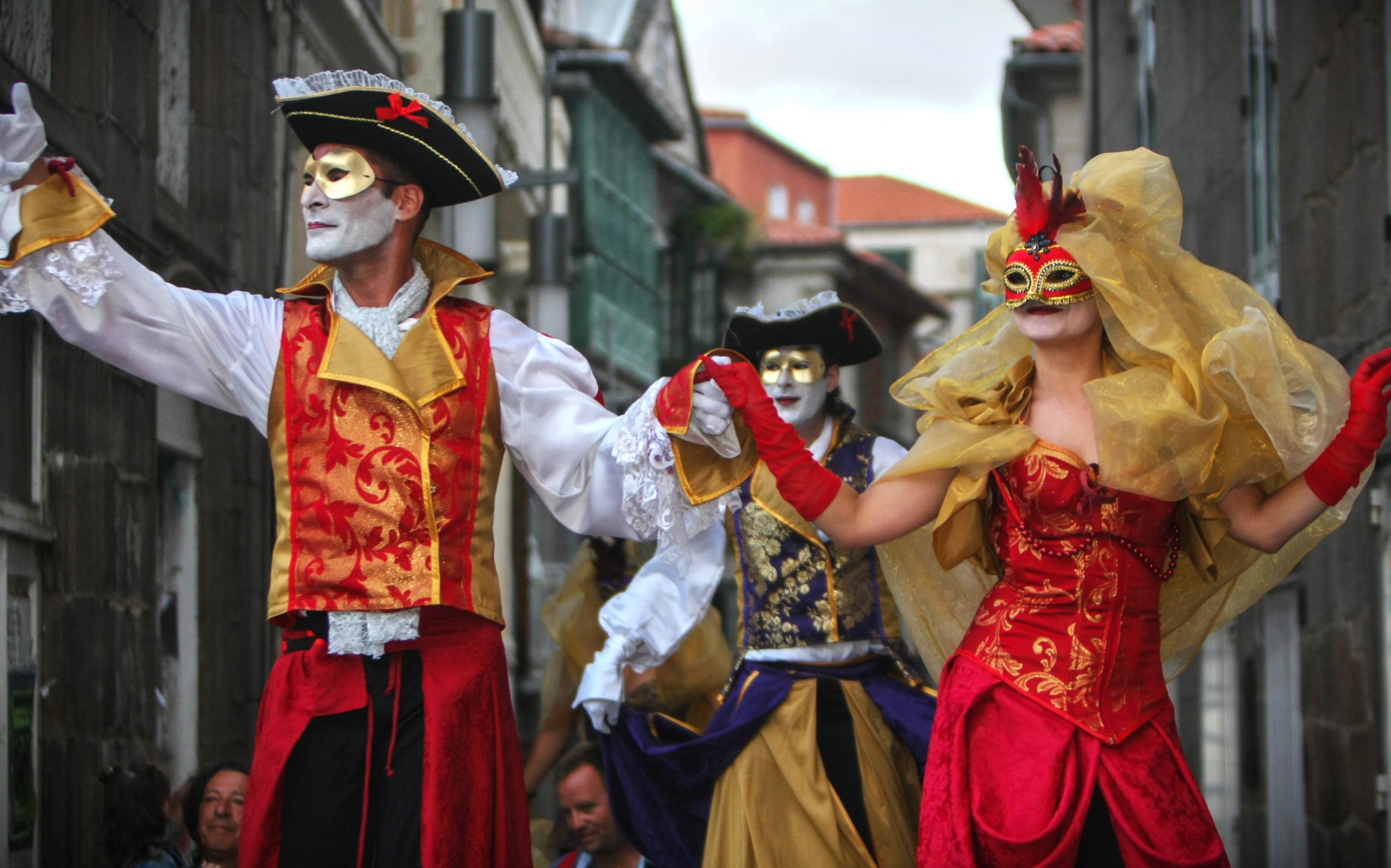Artistas sobre zancos desfilan de la mano con máscaras y ostentosos trajes en rojo y oro.