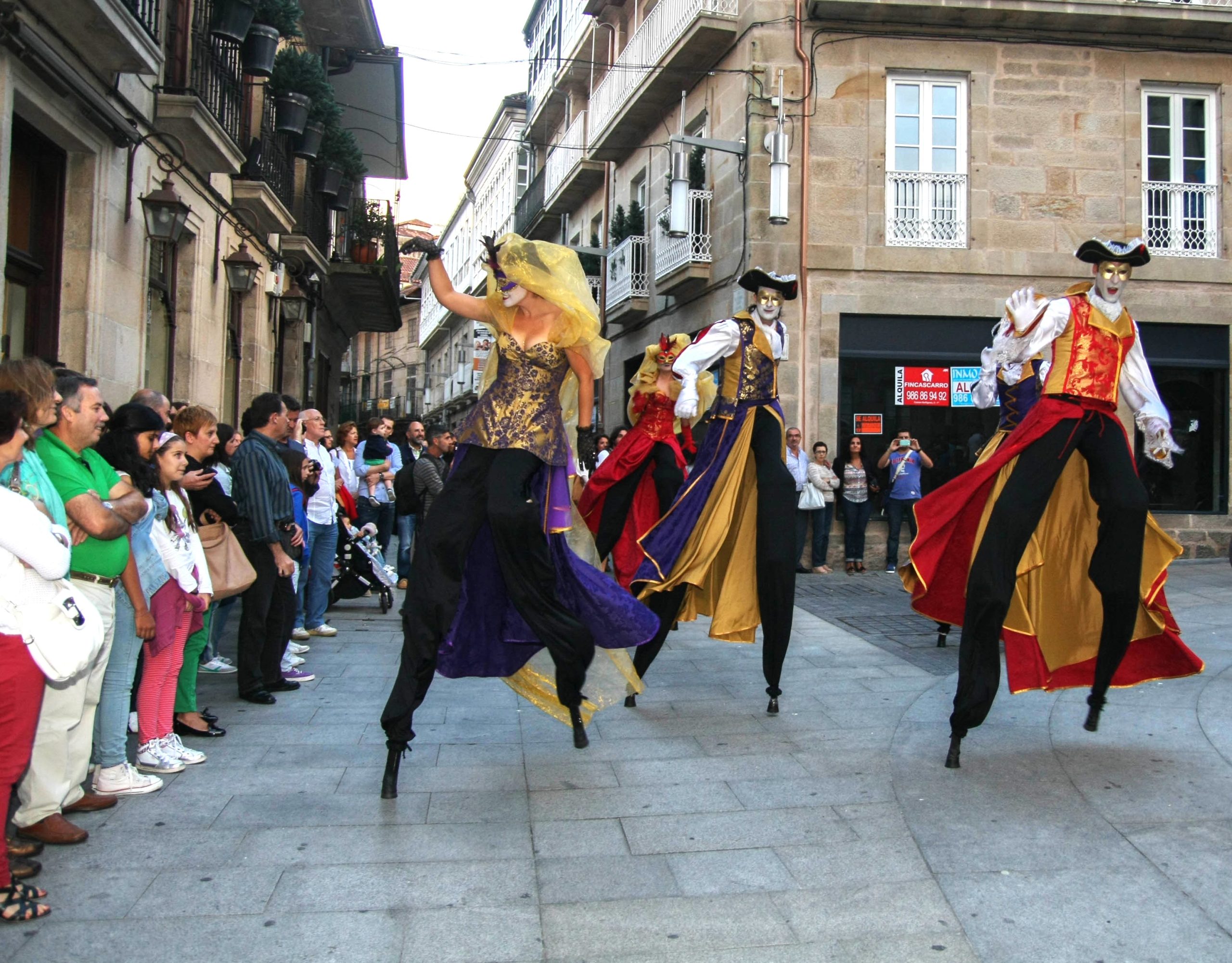Tres artistas con máscaras y trajes de gala venecianos danzan sobre zancos en una plaza de piedra.