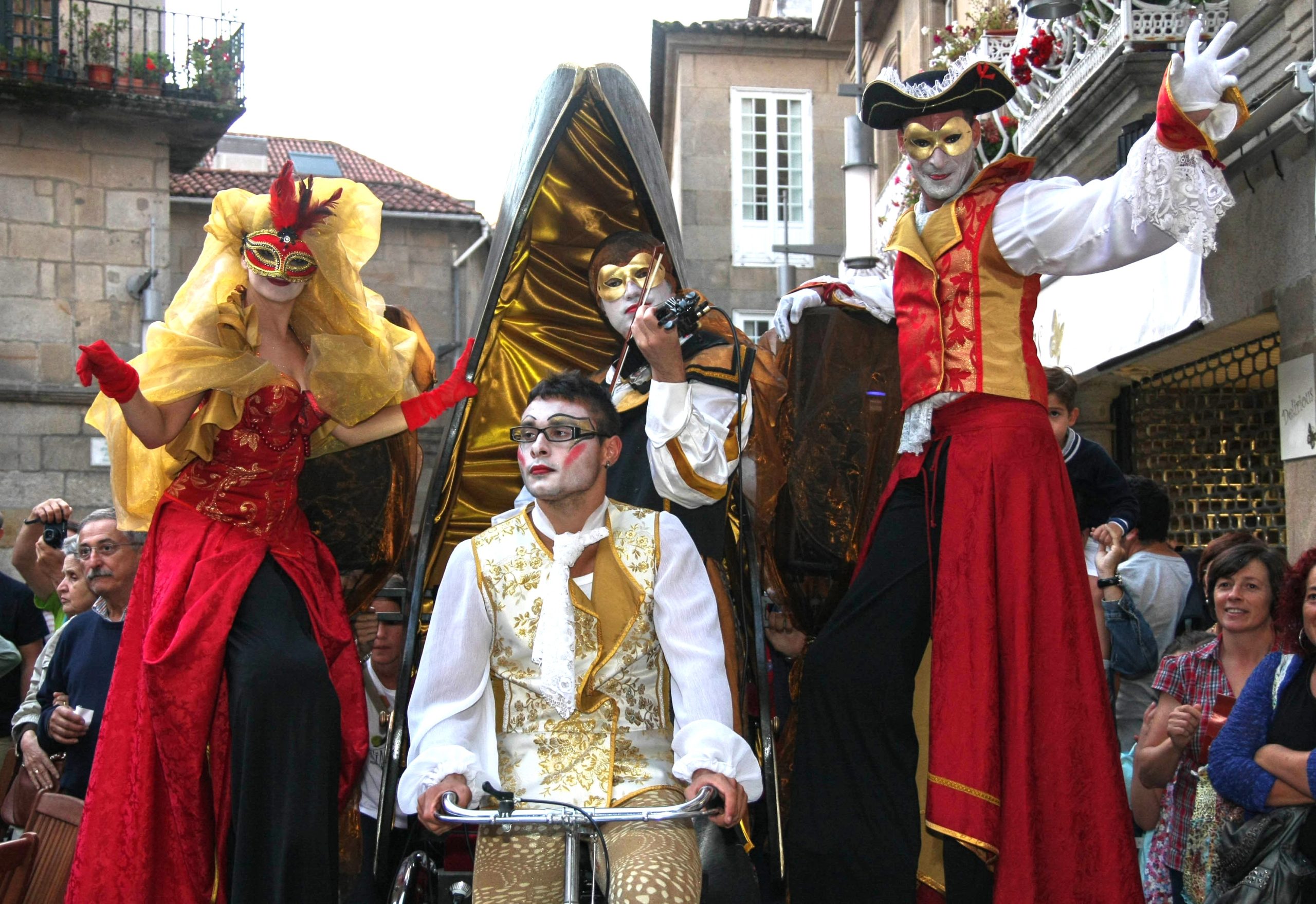 Un grupo de artistas con máscaras y trajes de gala desfila en una plaza.