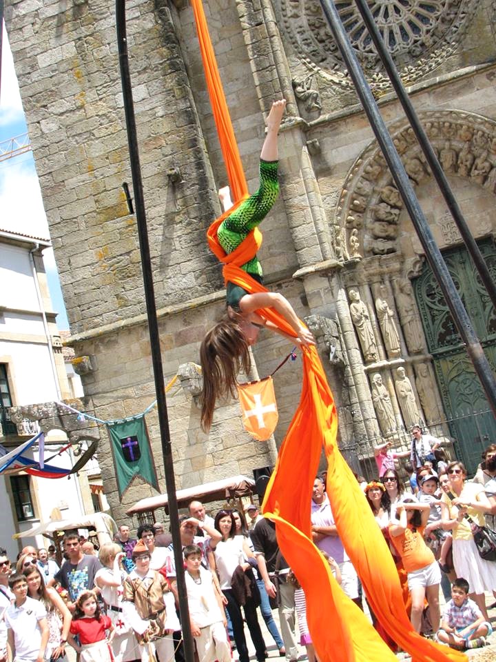 Acróbata con mallas verdes cuelga boca abajo de vibrantes telas naranjas frente a una catedral de piedra.