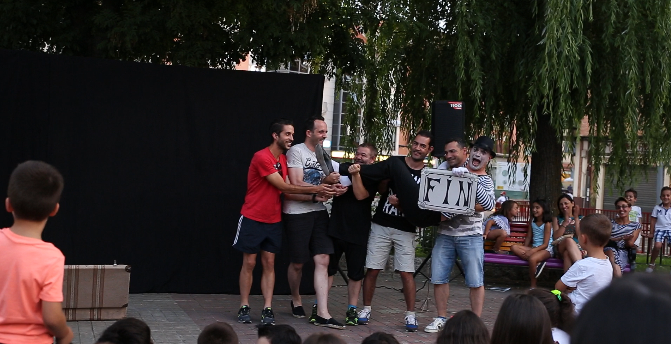 Un grupo de voluntarios sostiene al mimo en volandas mientras este, con una expresión cómica, alza un cartel con la palabra "FIN".