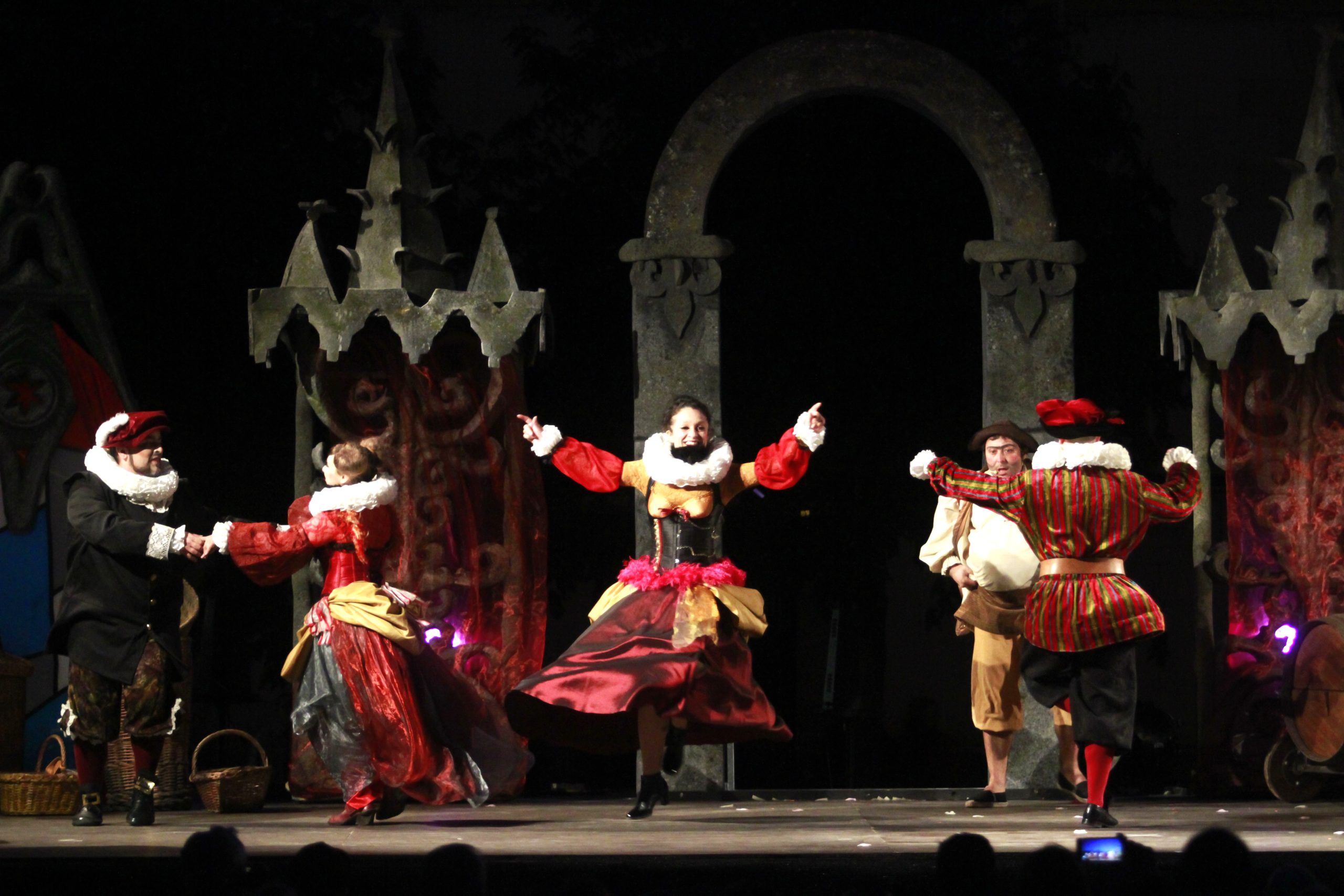 Cinco actores con trajes de época bailan animadamente en un escenario bajo un arco de piedra.