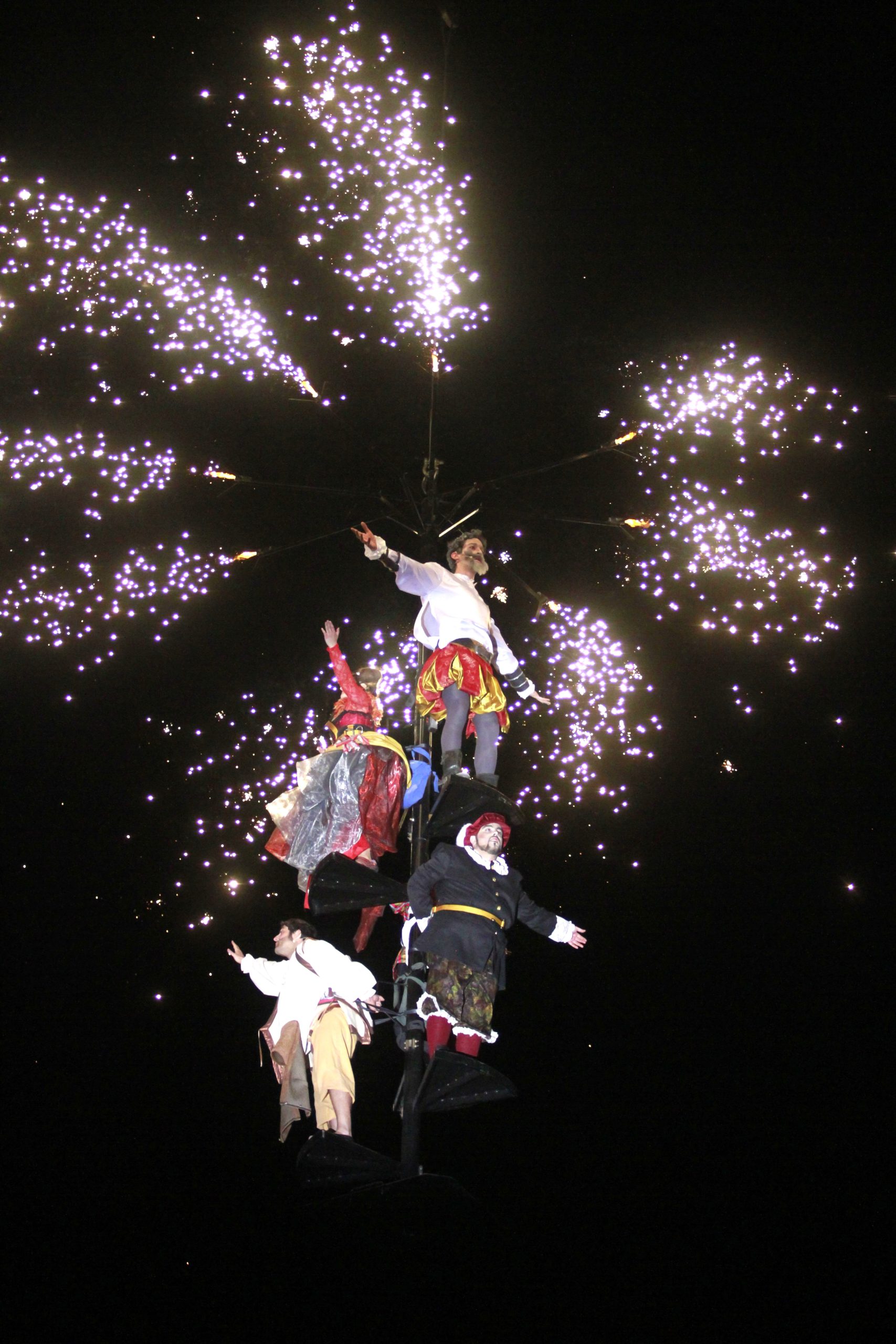 Don Quijote, Sancho Panza y dos actores con trajes de época están suspendidos en una estructura vertical contra el cielo nocturno.