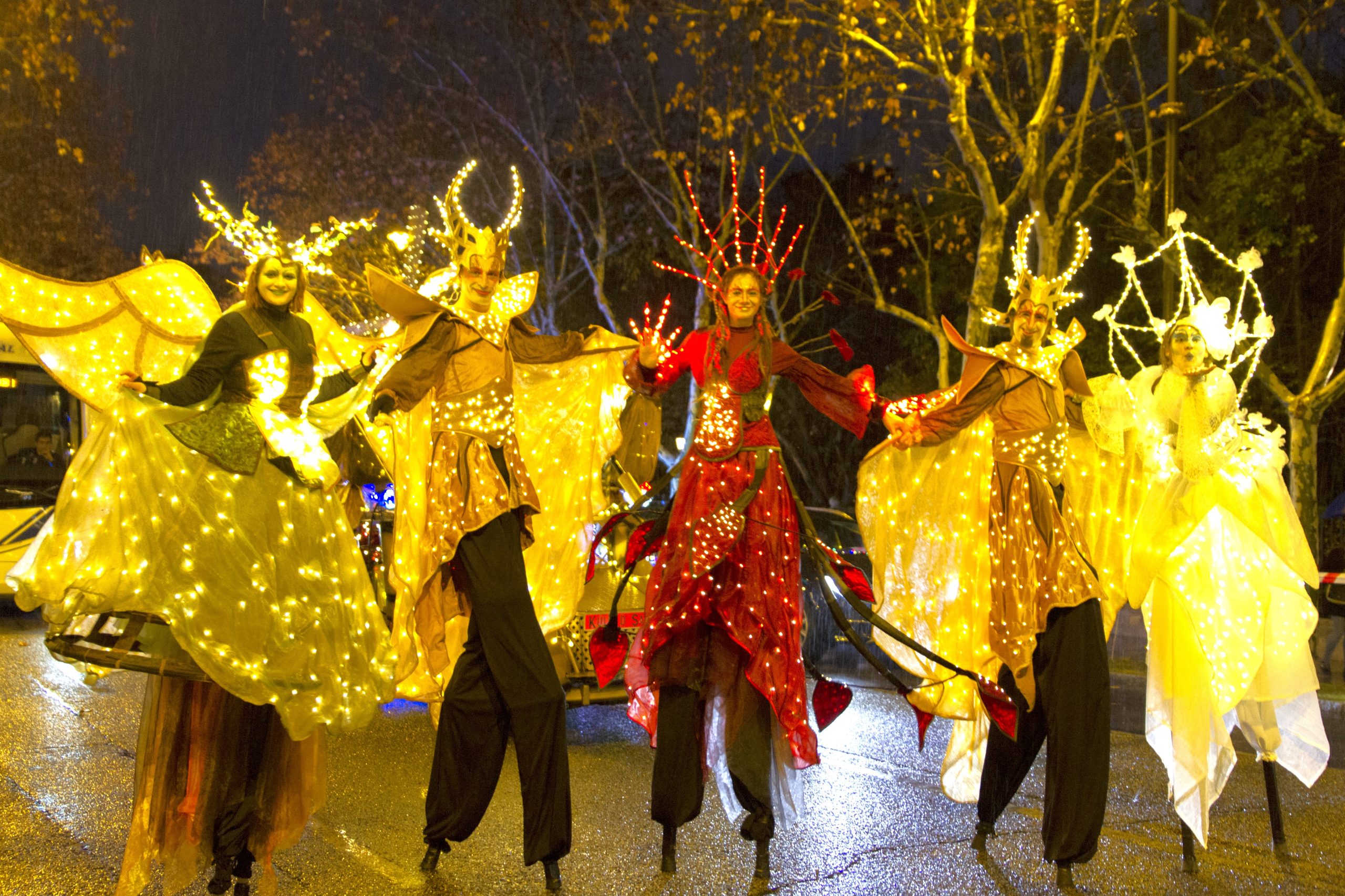 Cinco artistas sobre zancos visten trajes brillantes con luces LED amarillas y rojas.