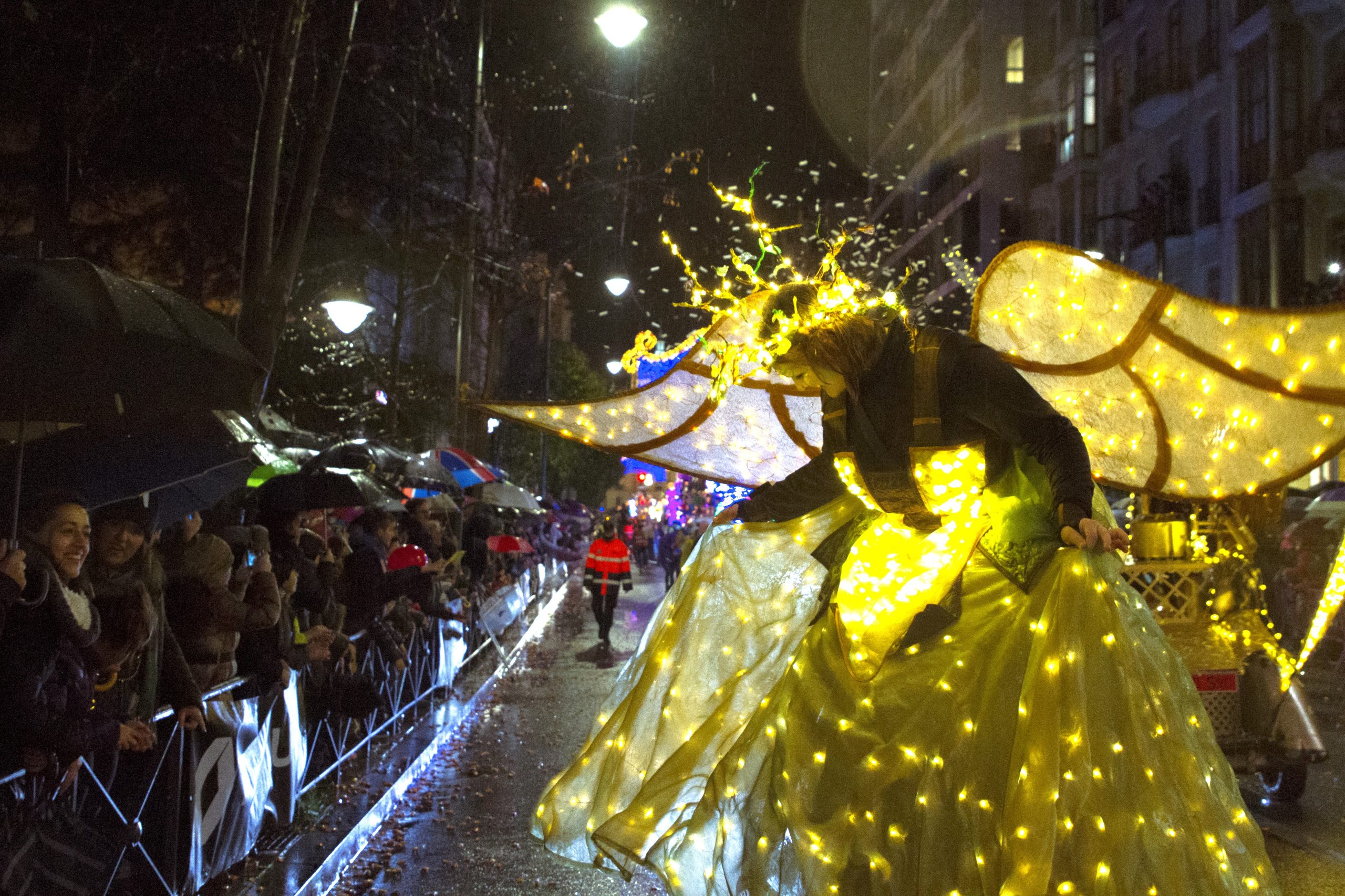 Actriz en zancos, con alas y vestido iluminados por cientos de luces LED desfila frente a una multitud