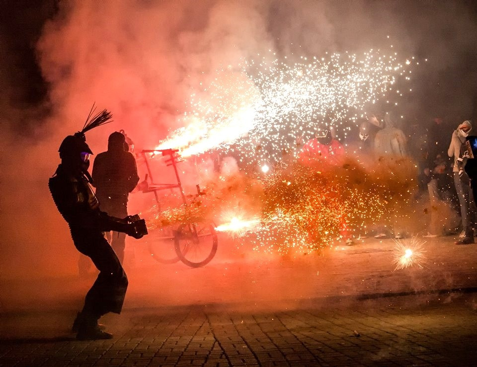 Un artista en silueta con casco de crestas dispara ráfagas de chispas naranjas de noche.