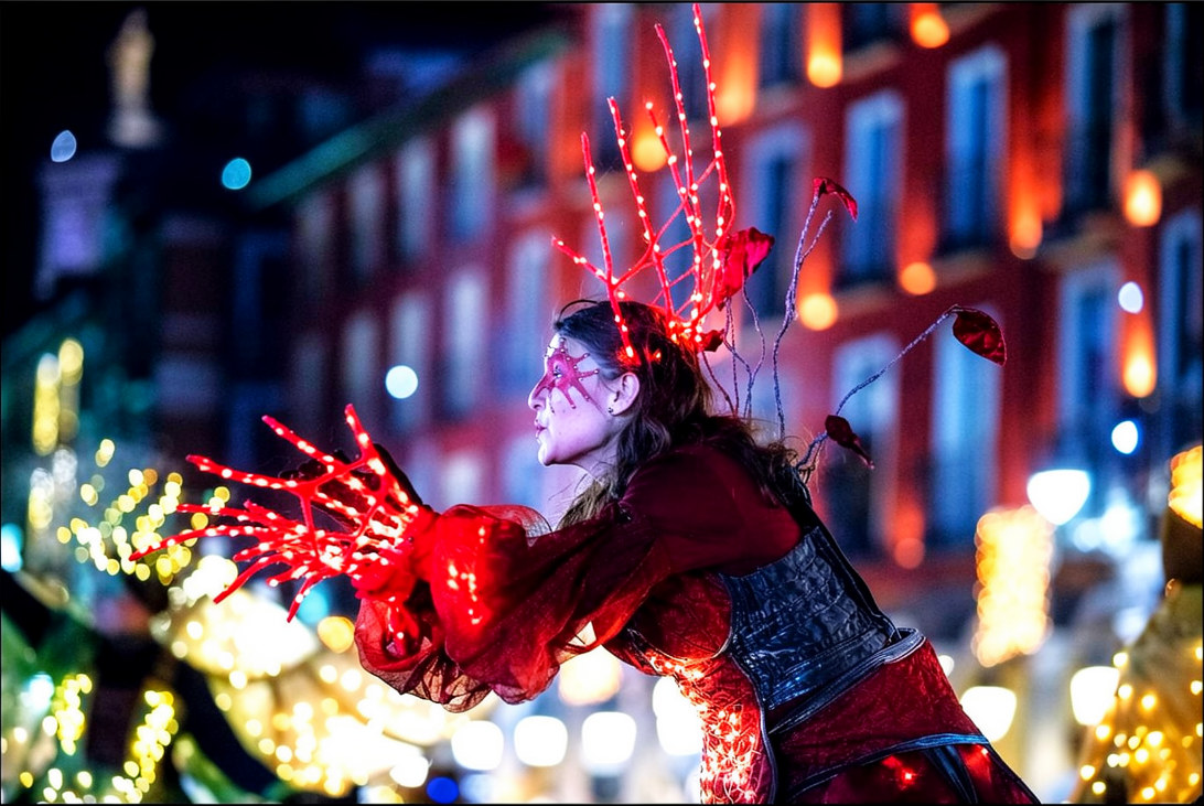 Actriz en zancos, con traje granate y un tocado de ramas rojas iluminadas con LED.